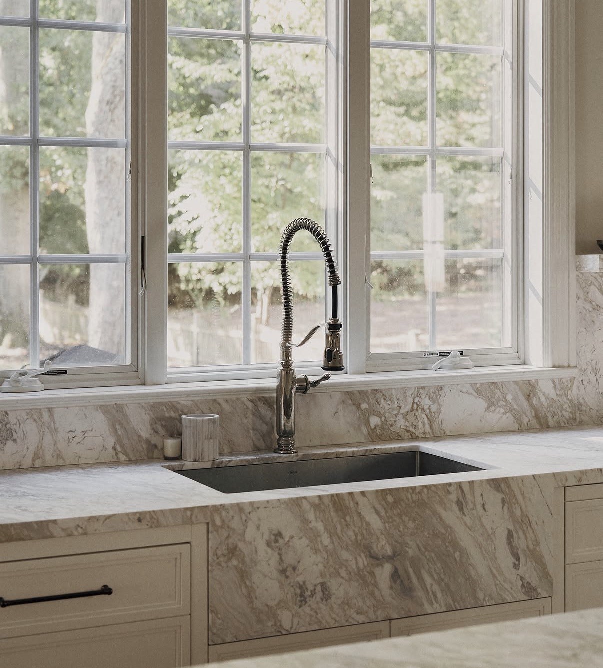 Stone apron-front sink and matching veined backsplash beneath kitchen windows